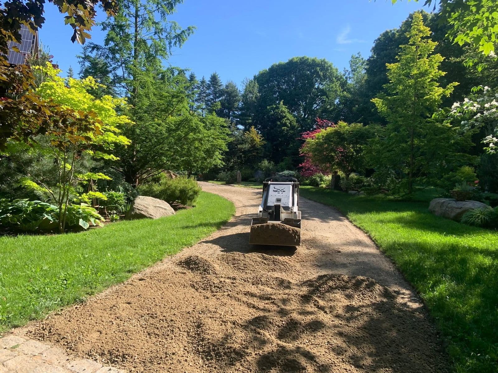 Compact track loader spreading gravel along a curved driveway between planted beds