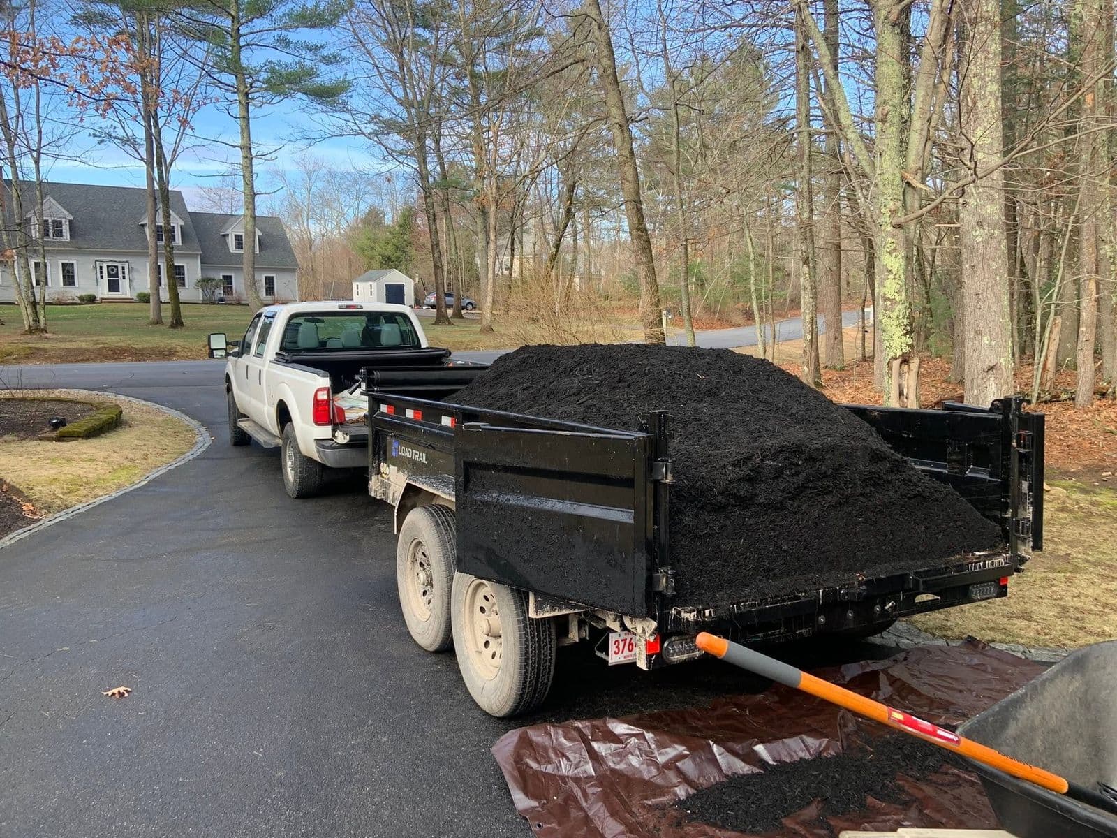 Black dump trailer full of dark mulch behind a pickup truck on a paved driveway