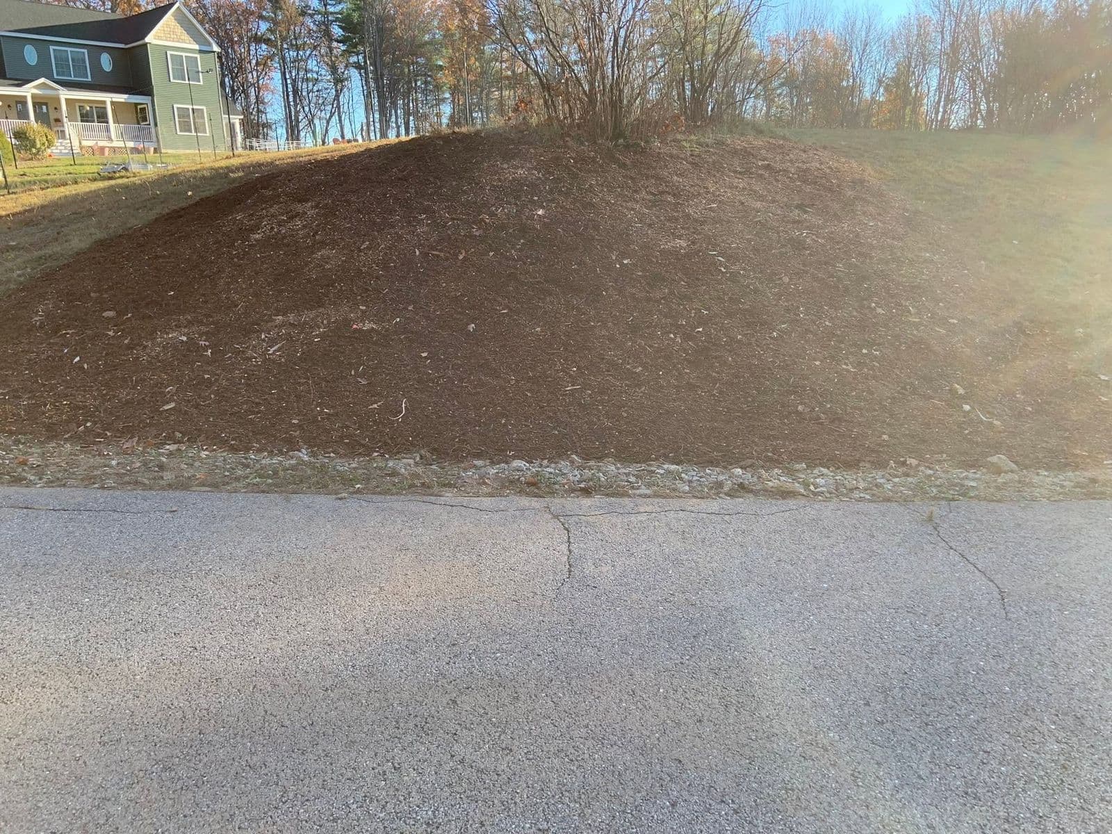 Mulched embankment beside a paved road with a house and bare autumn trees behind