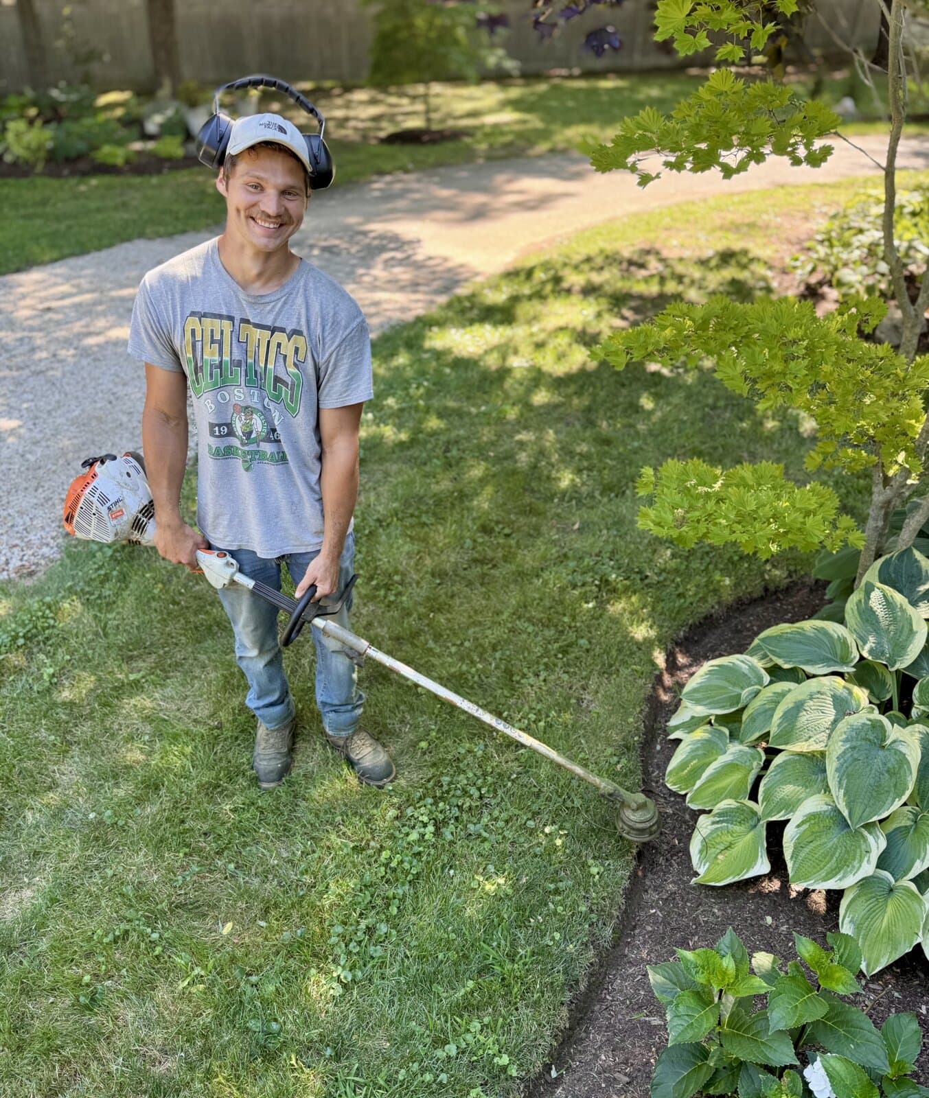 Ryan Burgess of Bellamy Landscape Services smiling on a client's lawn, holding a Stihl string trimmer beside a curved mulch bed of hostas